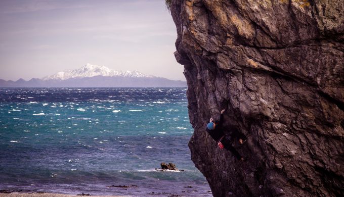 Man climbs steep rock with sea and mountain in background